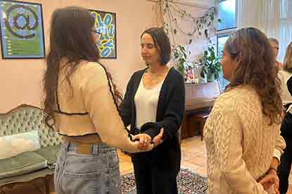 3 women standing. One is watching. One has her eyes closed and the other is holding her arms. Other people doing similar processes in the background
