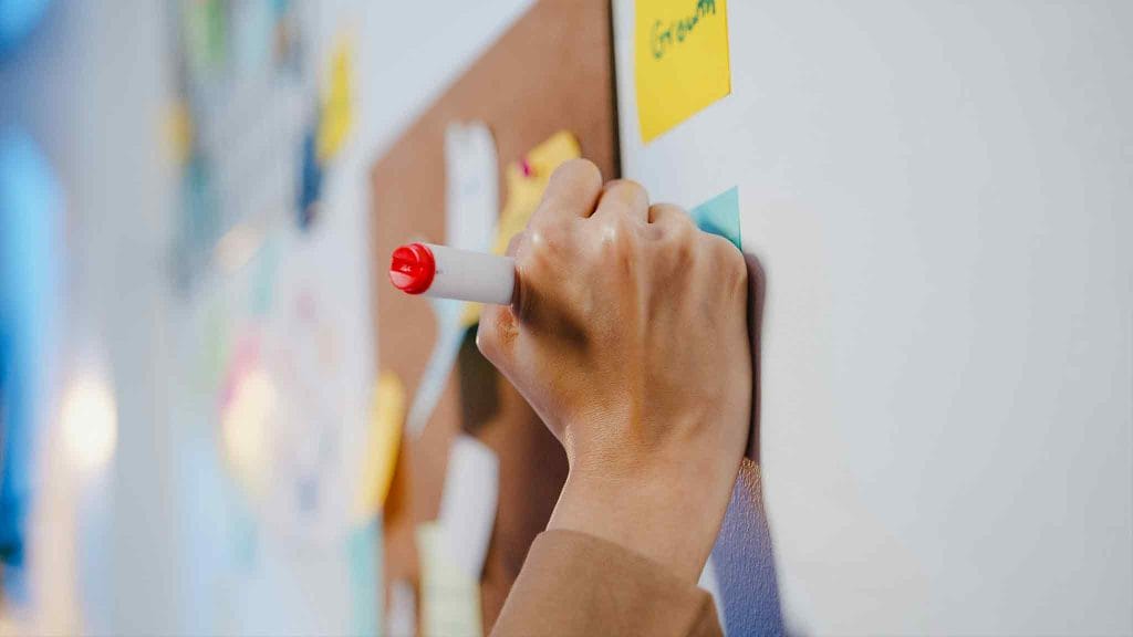 someones hand writing on a post it notes on a whiteboard with other coloured postit notes on it