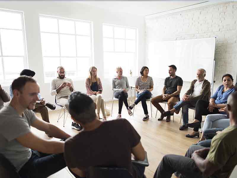 group of people sitting on chairs in a circle in white room with large windows